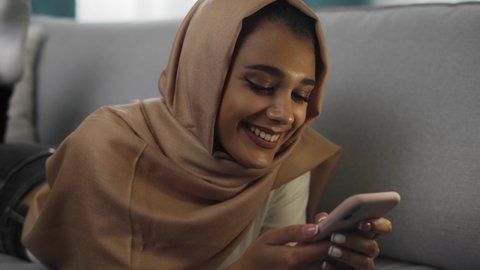 Charming young Indian woman with beautiful smile, wearing hijab scarf is taking rest on couch, lying relaxed, using phone, communicating in online chat, browsing social networks.