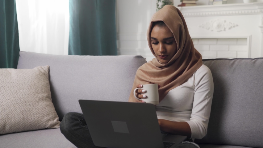 Indian woman in hijab scarf sits on couch, using laptop, busy working online, typing, searching the Internet, drinking coffee or tea. Muslim lady freelancer doing remote job. Royalty-Free Stock Footage #1060434031