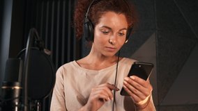 Portrait of beautiful singer in headphones looking text of new song in smartphone working in modern sound recording studio. Music production