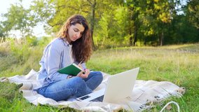 Pretty young woman in casual clothing sitting on blanket at green park and using modern computer studying. Female student with dark hair writing in notebook outdoors.