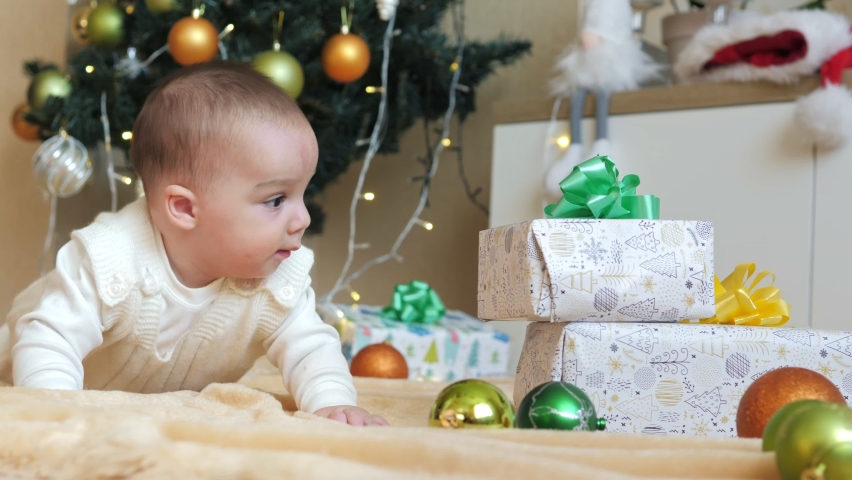 Little baby boy lying on a cozy blanket near the Christmas tree and garlands in the house next to the gifts