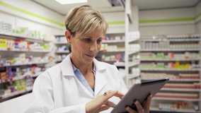 Female pharmacist adjusting settings on digital tablet while assistant sorts out scripts behind counter in pharmacy 