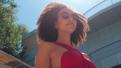 Curly haired African millennial woman moves in sun loght, walks with graceful slow gait past camera, adjusts fashionable glasses her hand, sunbathes in open air blue clean sky, shooting from below