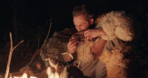 Primitive hunter gather wearing sheepskin squatting eating meat by the light of the flames of a fire at night tearing the meat from the bone with his teeth - wild caveman