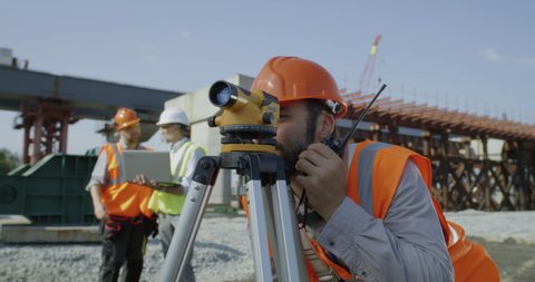 Bearded contractor speaking on radio and gesturing thumb up while using theodolite then calling colleagues to check measurements during work on construction site