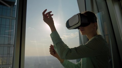 Woman using virtual reality headset and looking around against view on cityscape from panoramic skyscraper window in office. Sun lens flare. VR, opportunity, sightseeing, discover, technology concept
