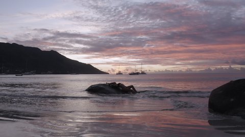 A dramatic sunset, a girl lies on a rock, ocean waves gently wash her body. Seychelles slow motion