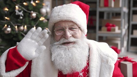 Funny old bearded Santa Claus, Saint Nicholas talking to camera greeting on Merry Christmas Happy New Year, standing in warehouse workshop, video calling by webcam online virtual chat concept.
