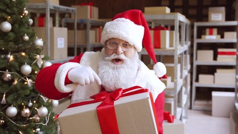 Funny old bearded Santa Claus, Saint Nicholas holding present, talking, giving gift box to camera greeting on Merry Christmas Happy New Year, standing in warehouse workshop, xmas sale delivery concept
