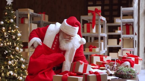 Old funny Santa Claus, Saint Nicholas packing presents gift boxes in sack bag preparing post shipping fast xmas delivery parcels walking in workshop. Merry Christmas shipping delivery concept.