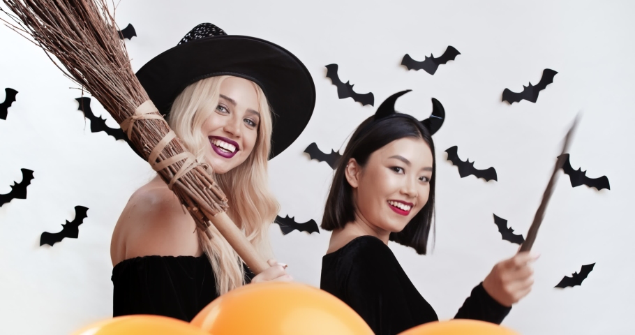 Halloween enchantresses. Two multiethnic women in with costumes smiling and flirting at camera, blonde holding broom, asian playing with magic wand, studio background