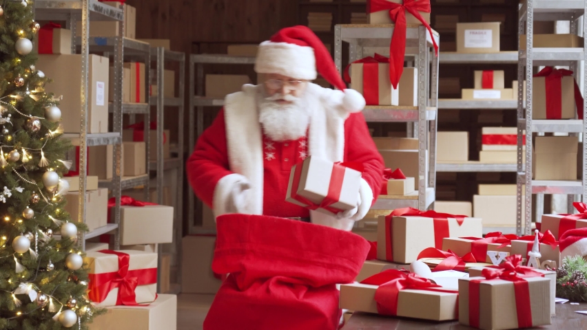Old funny Santa Claus, Saint Nicholas packing presents gift boxes in sack bag preparing post shipping fast xmas delivery parcels walking in workshop. Merry Christmas shipping delivery concept. Royalty-Free Stock Footage #1060450681
