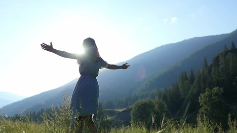 A young girl in a beautiful dress whirls happily in slow motion in a field against a background of mountains. Beautiful carefree woman enjoying nature and sunrise sunlight