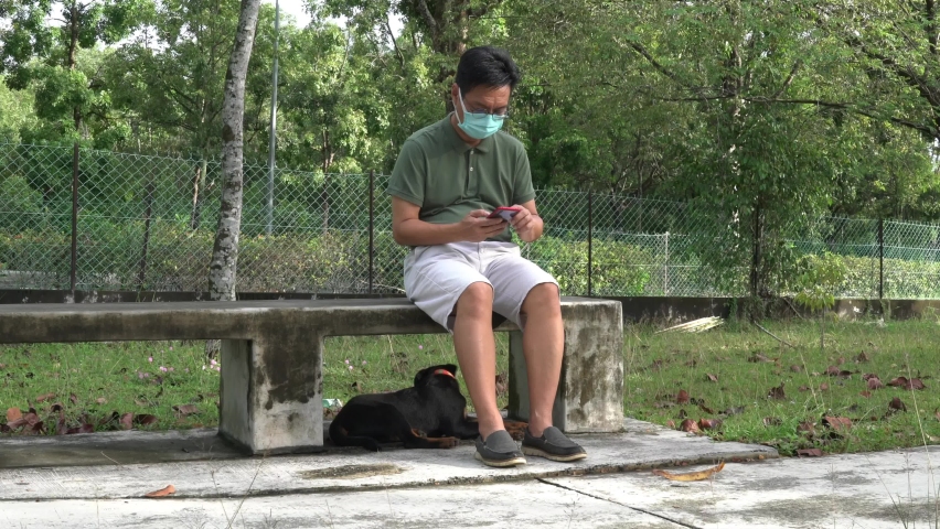 Man wearing face mask sitting on a bench and reading his cellphone, with puppy dog at his feet. Time with pet dog in the public during pandemic concept.