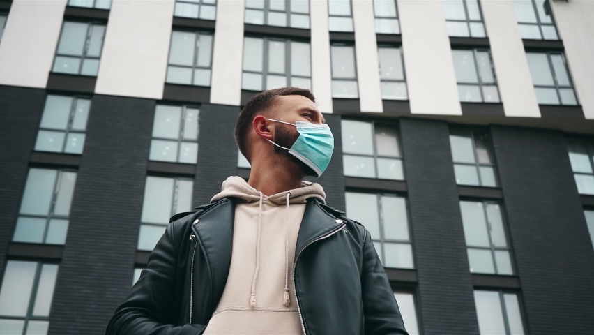 Young stylish man wearing a protective mask looking around before modern building