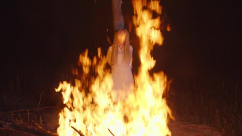 Screaming helpless attractive woman in white dress, convicted as witch, condemned to death by burning for heresy and witchcraft, executing on stake outdoors at night during medieval witch hunt.