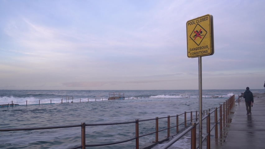 A yellow sign with an inscription that the pool is closed - dangerous conditions is standing alongside the rock pool by the ocean. Lonely silhouette figure is moving away from the pool.
