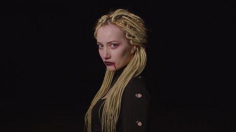 Creepy bloodthirsty charming woman vampire with dreadlocks and bloody pale face spitting fresh blood on ground after successful hunt on halloween night, looking dangerous and horrible.