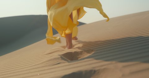 Fashionable shot: close up view of slim woman legs walking by the rippled sand dune surface. Female model in bright yellow flattering dress walking toward horizon. 4K stylish clothes is waving on wind