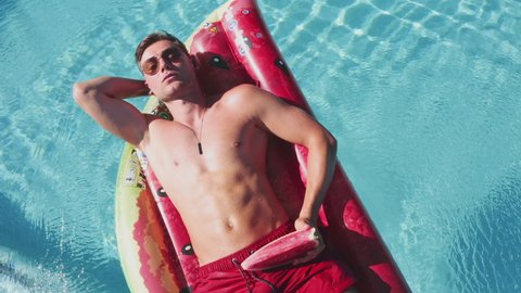 Drone Overhead Wide Zoom Out Of Young Muscular Man Relaxing On Inflatable Watermelon In Swimming Pool