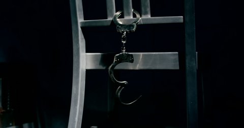 Slow motion medium tracking shot of a pair of handcuffs dangling from the back of a chair. One cuff is unlocked.