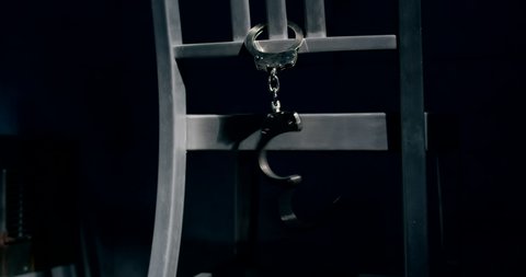Medium tracking shot of a pair of handcuffs dangling from the back of a chair. One handcuff is unlocked and swinging.