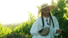 Young woman walks in white dress and straw hat in the vineyard at sunset. Slow motion video 4K