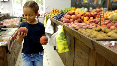Little kid girl with two peaches in hands is walking along grocery rows toward to cashier, steadicam shot.