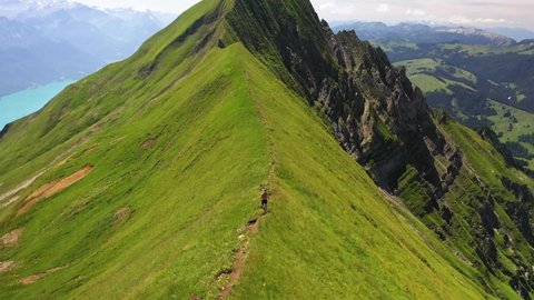 Drone shot following a running young, caucasian man on top of Hardergrat ridge near Interlaken in Switzerland. Majestic and massive mountain with a steep slope on each side with green fields.