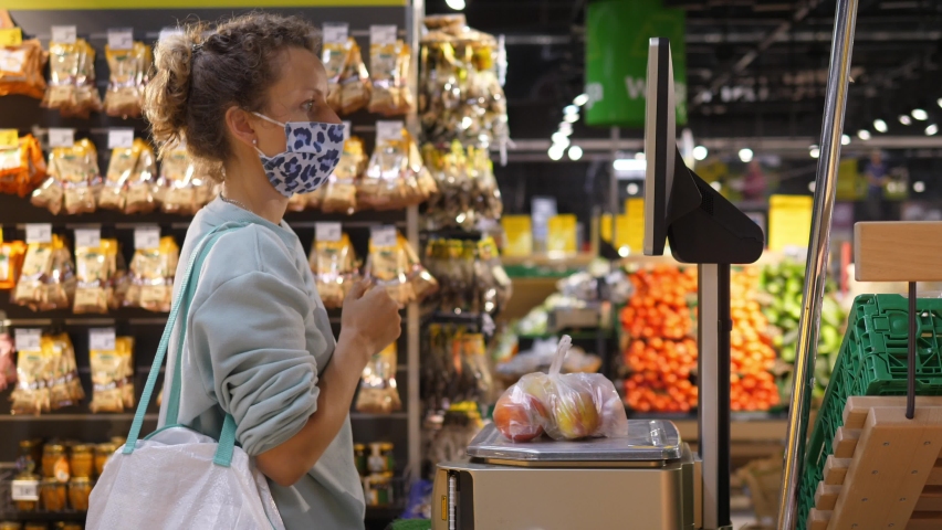 Side view of a blonde girl in a face mask weighing fruits on an electronic scale in a supermarket. Shopping during covid-19 pandemic Royalty-Free Stock Footage #1060492567