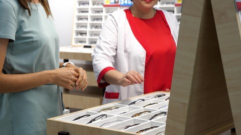 Professional female optician helping client to choose a spectacles in optical store. Young woman is choosing eyesight glasses.