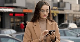 Thoughtful young lady wearing beige coat and eyeglasses standing on street, holding smartphone and looking around. Concept of people and technology.