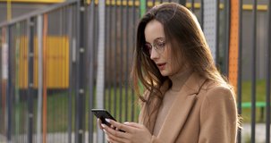 Attractive young brunette in eyewear and beige coat feeling happy while looking at smartphone screen on fresh air. Concept of city lifestyle and modern technology.