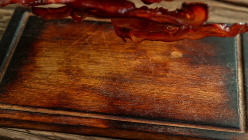 Super Slow Motion Shot of Roasted Bacon Slices Falling on Wooden Board at 1000 fps. Royalty-Free Stock Footage #1060495804