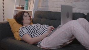 Young pregnant woman is lying on the sofa talking to her husband using computer at home. The pregnant woman communicates with her husband via video chat on the computer.