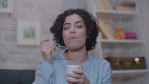 A cheerful young woman is sitting on an armchair at home and eating yogurt.