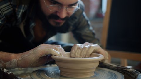 Slow motion close-up of young professional potter making bowl from clay on throwing wheel concentrated on creative work. People and job concept.