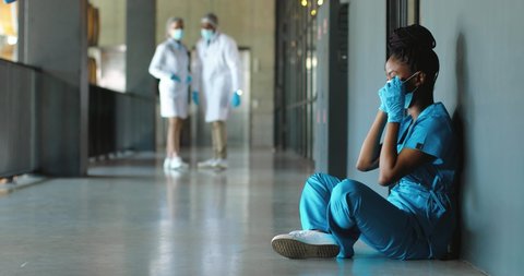 Tired sad African American woman doctor in uniform and gloves taking off medical mask and leaning on wall in despair. Upset female nurse resting, having break. Coronavirus. Rest at work. Covid-19.