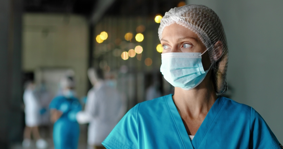 Portrait of tired Caucasian pretty woman doctor in hat and medical mask looking at camera. Close up of female physician. Medic or nurse in clinic. Indoor. Coronavirus concept. Protected. Royalty-Free Stock Footage #1060503742