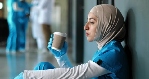 Tired Arab young woman doctor in hijab and gloves drinking coffee and leaning on wall sitting on floor. Muslim female nurse in headscarf resting, having break, sipping drink. Coronavirus work.