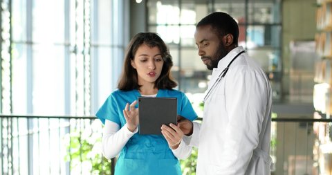 Mixed-races young couple of male and female doctors standing in hospital, talking and using tablet device. Multi ethnic man and woman, physicians discussing work and watching on gadget.