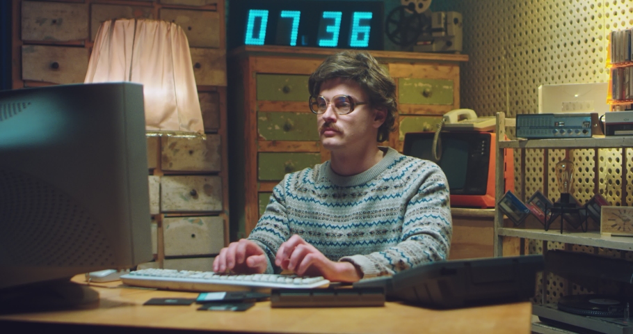 Caucasian male nerd in glasses with mustache sitting at desk in retro room and working on computer. Man programist typing on keyboard while studying. Vintage style of 90's. Hacker from 80's. Royalty-Free Stock Footage #1060503877