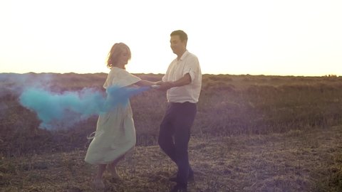 Gender reveal party with caucasian couple who spin round holding hands with smoke bomb in Slowmo. Man and pregnant woman are expecting baby boy. They are dancing and smiling in summer field on sunset.