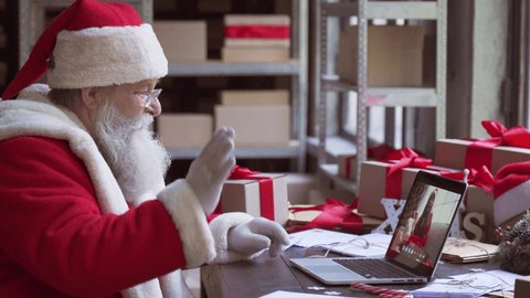 Santa Claus video calling kid talking to kid waving hand greeting on Merry Christmas, Happy New Year in virtual video online chat meeting on laptop sitting at workshop table sending gift on xmas eve.