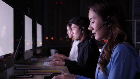 3 pepople, Late night shift call center business concept. Beautiful female Asian customer service on headphone, working, smiling, talking, pleased to service, among blurry bacground of coleagues