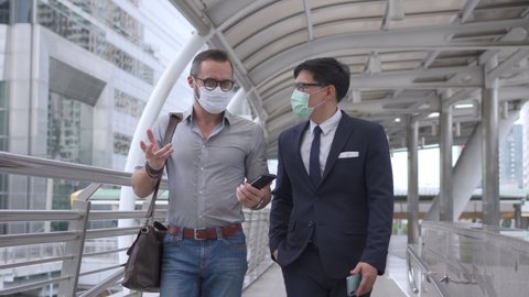 Business people Caucasian with Asian man office worker colleague wearing face mask for protect covid-19 virus walking together on office district and discussing business project with using smartphone.