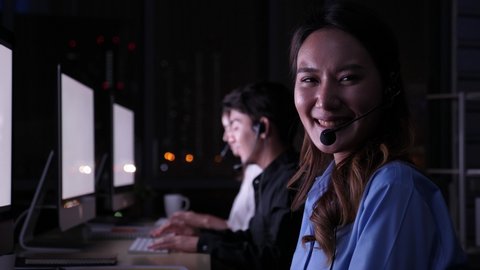 Selective focus of beautiful asian call center work with white screen desktop at night, talk to headphone, look at screen and turn to camera to smile and nod gentley with the background of colleagues