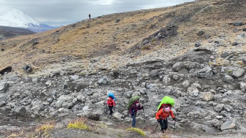 Three young women travelers with backpacks and trekking poles are walking on the stones. Trekking in Klyuchevskoy volcano park. Travel to the Kamchatka Peninsula.