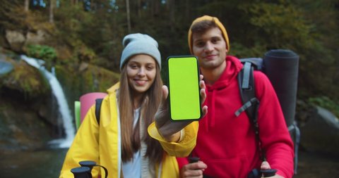 Portrait of happy Caucasian couple with backpacks standing in mountains and showing smartphone with green screen to camera. Woman and man near waterfall in forest holds cellphone with chroma key
