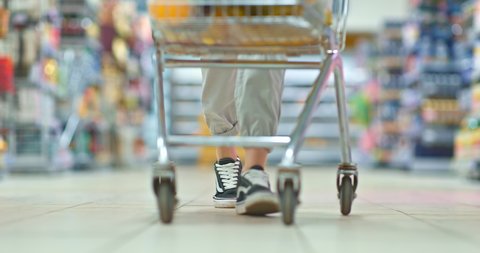 Close-up of a grocery cart driving through the supermarket. Women's feet walk through the supermarket and pull the cart.
